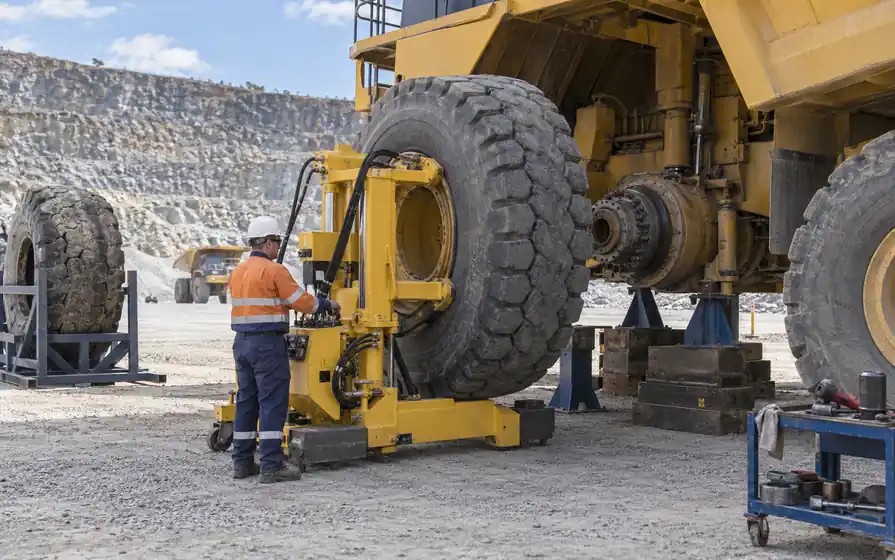 Operator aligning a huge quarry hauler wheel with a hydraulic tyre handler beside a supported exposed hub.